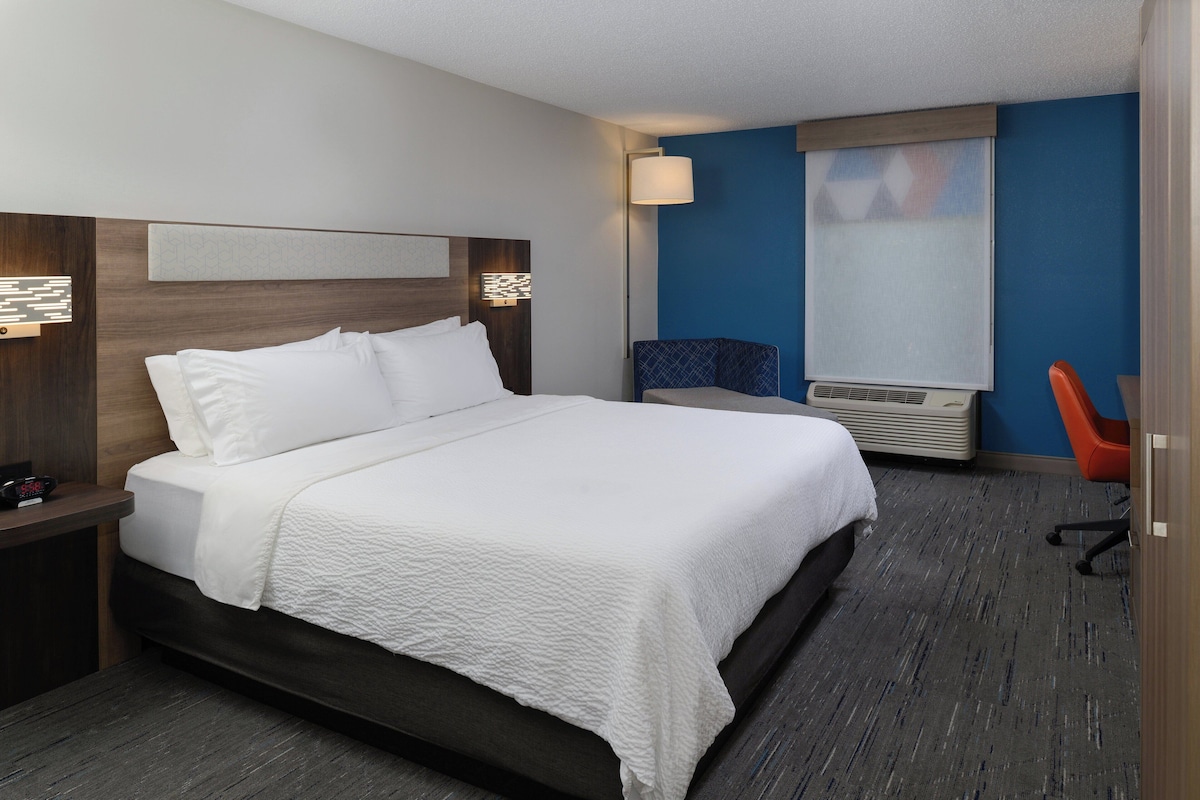 Une chambre d’hôtel avec un grand lit, un bureau, une chaise et une fenêtre avec des stores.