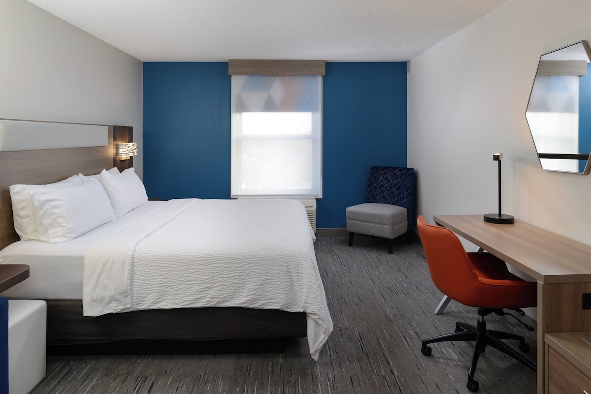 Habitación de hotel con cama, escritorio, una silla naranja, un espejo grande y ventana con persianas.