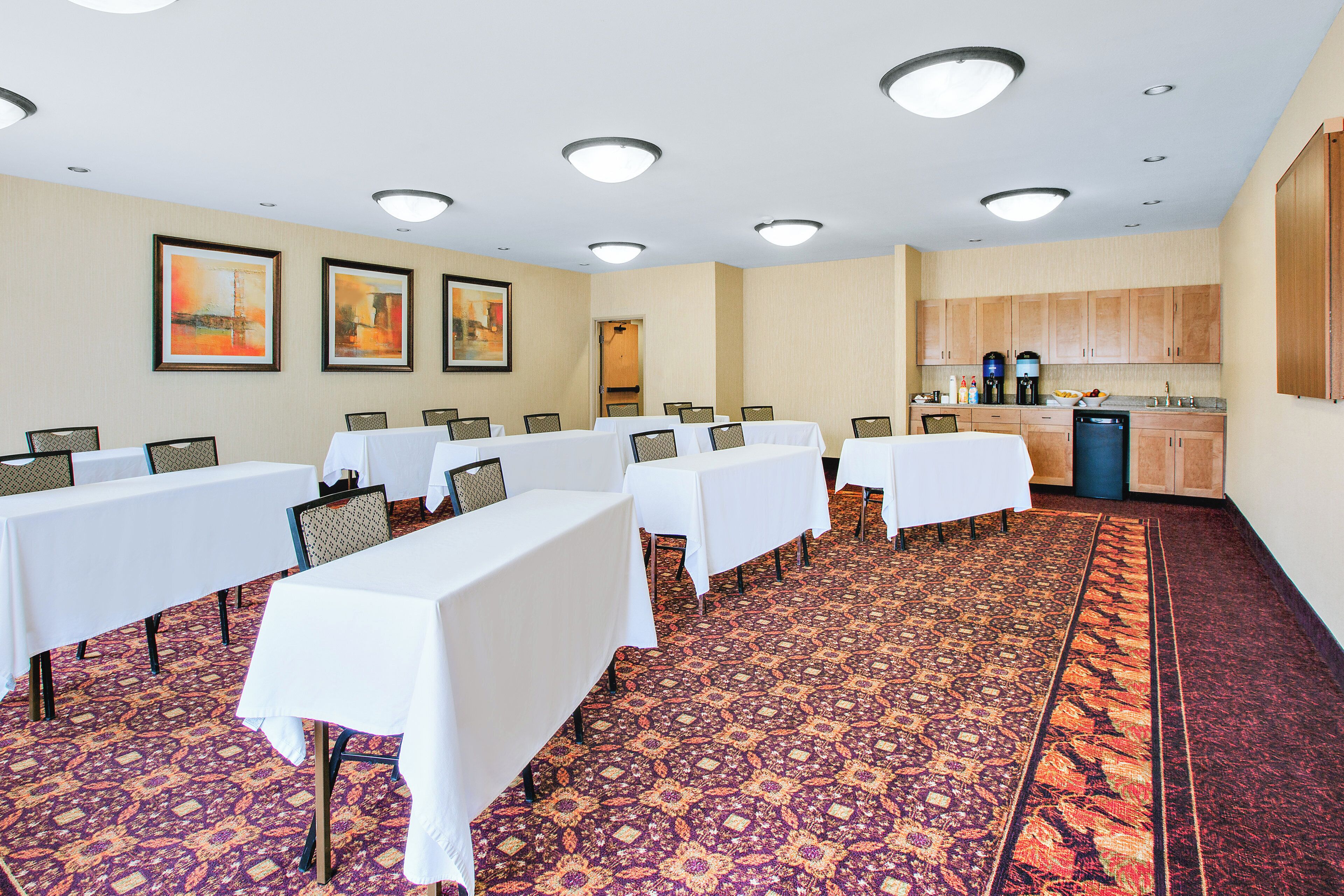 Un salón de conferencias con varias mesas y sillas, una kitchenette y suelo cubierto de alfombra estampada.