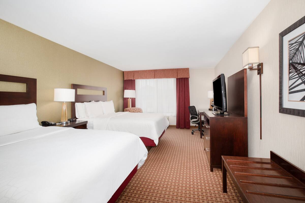 A hotel room with two beds, a desk, a TV, and a large window with curtains.