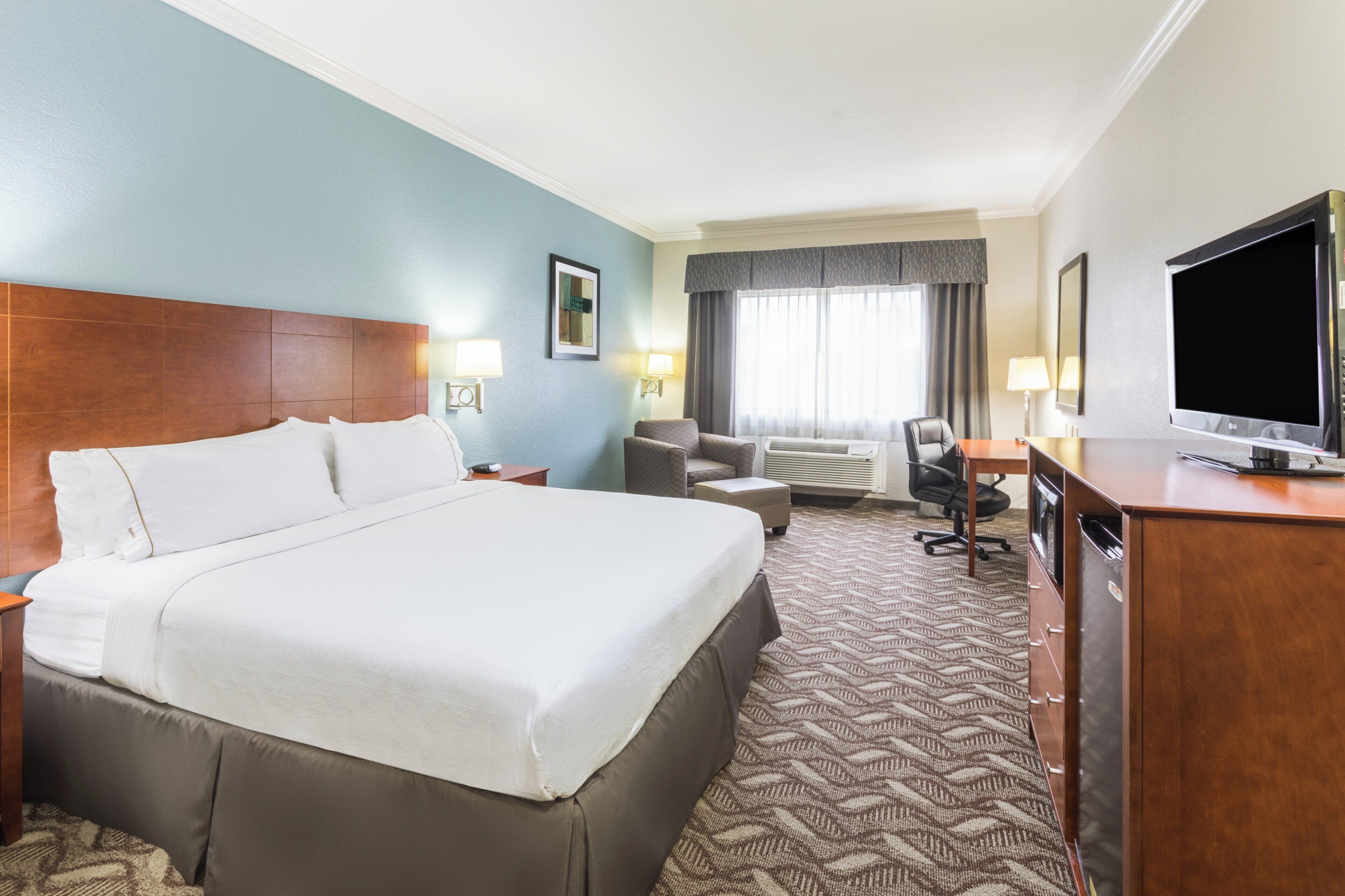 Une chambre d’hôtel avec un grand lit, un bureau, une chaise, une télévision et une fenêtre avec des rideaux.