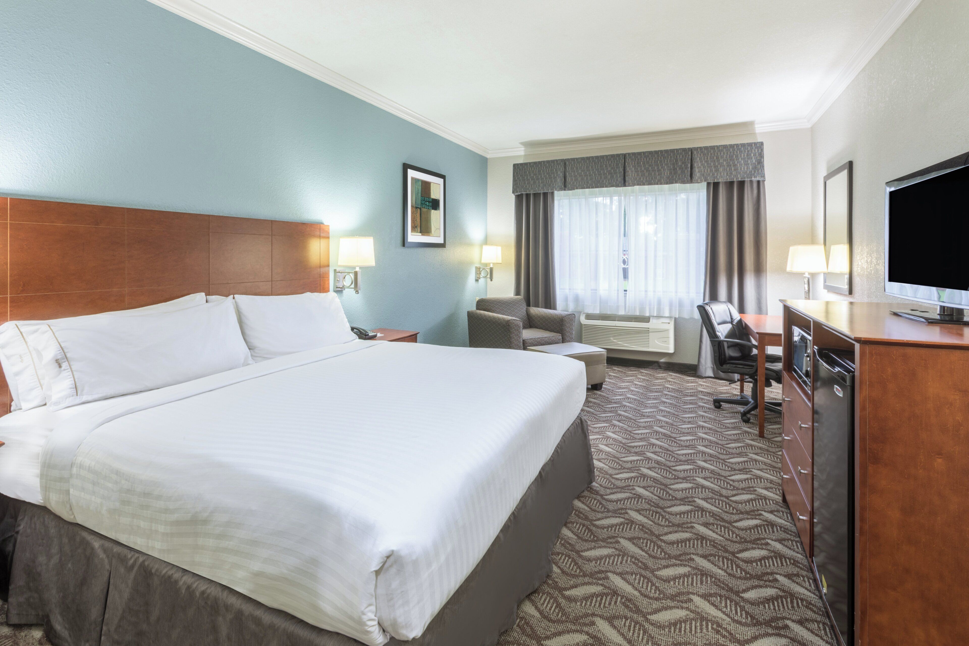 A hotel room with a large bed, a desk with a computer, a chair, a television, and a window with curtains.