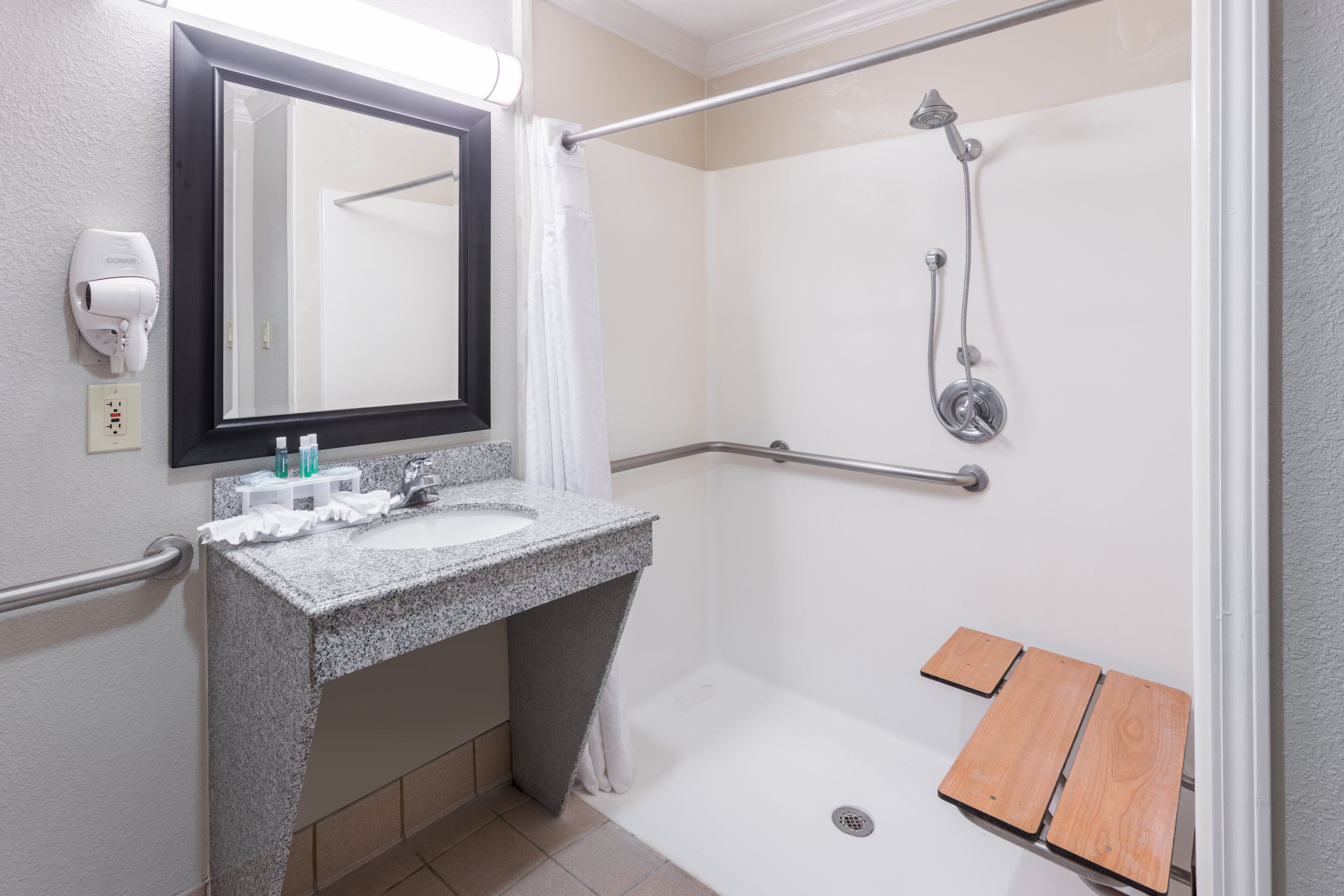 A bathroom with a granite sink, a mirror, a handheld showerhead, and a wooden bench.