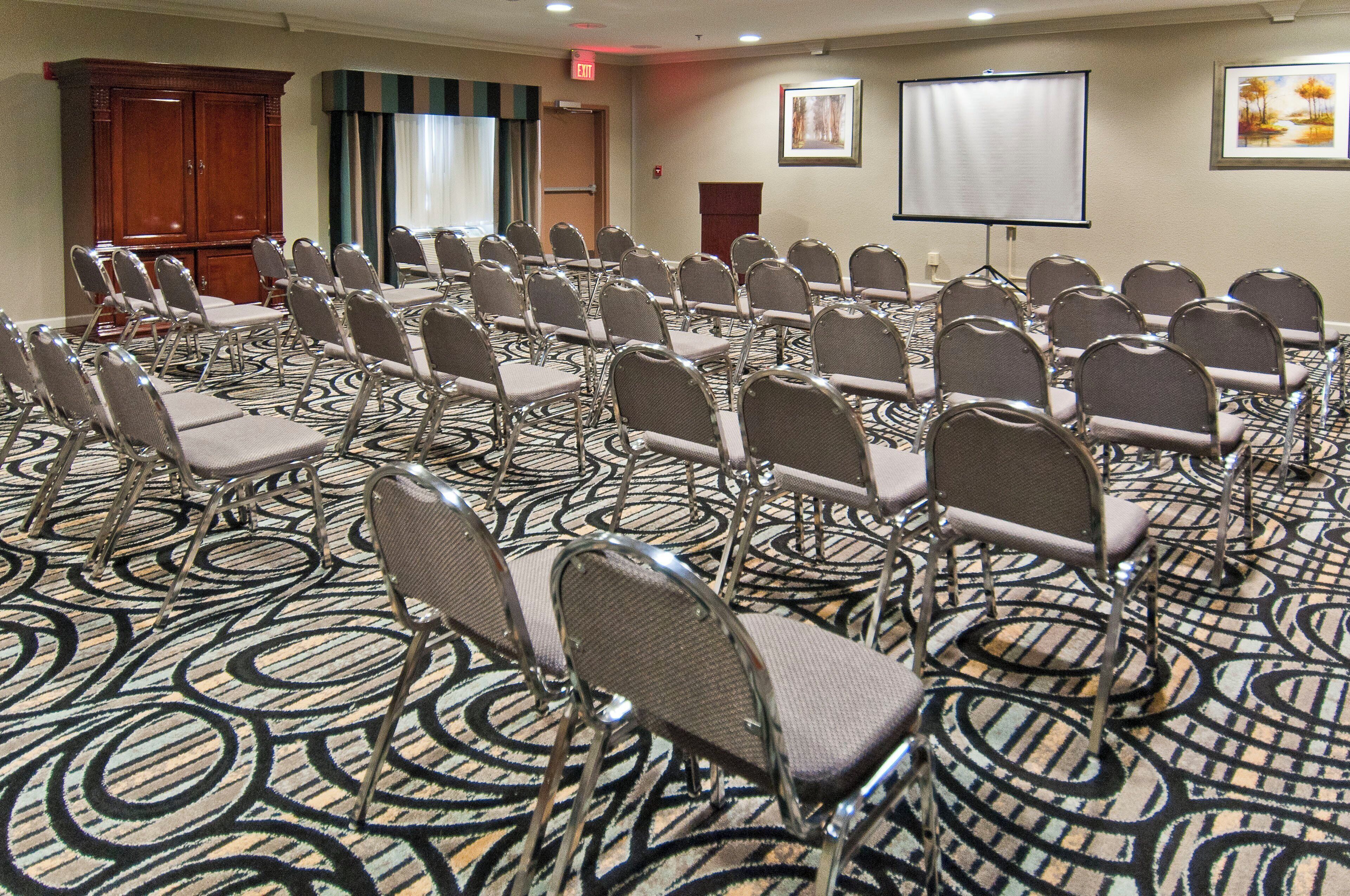 Un salón de conferencias con hileras de sillas, una pantalla de proyector y una alfombra con diseño circular.