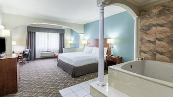 Habitación de hotel con una cama grande, un escritorio, una silla, un televisor, un baño con bañera y un ventanal con cortinas.