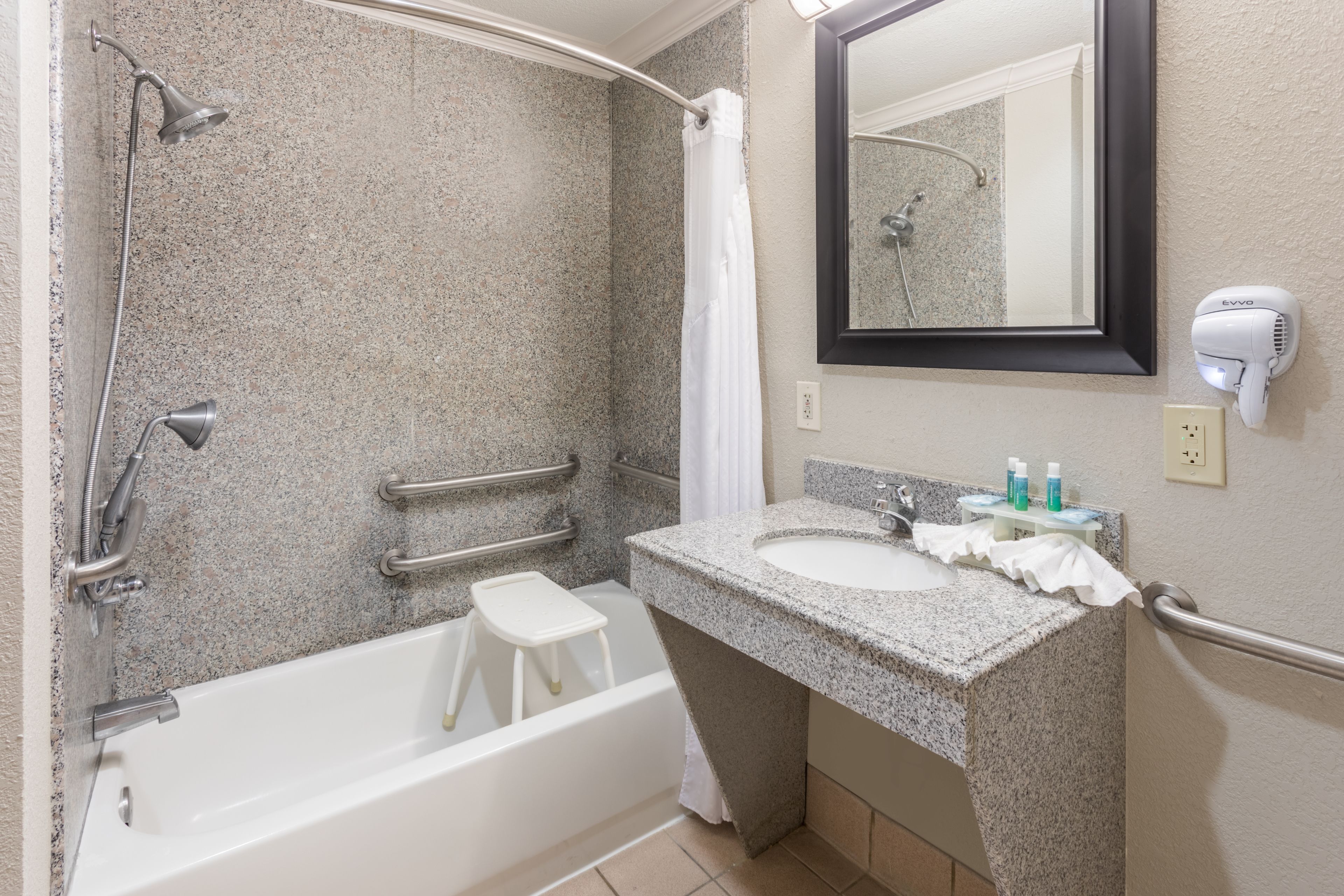 Un baño moderno con lavamanos de granito, ducha de mano, bañera blanca, un taburete, toallero y secador de pelo montado en la pared.