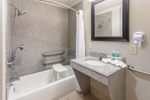 A modern bathroom with a granite countertop sink, a handheld showerhead, a white bathtub, a step stool, a towel rack, and a hairdryer mounted on the wall. - Holiday Inn Express Hotel & Suites Lake Charles by IHG (Lake Charles)