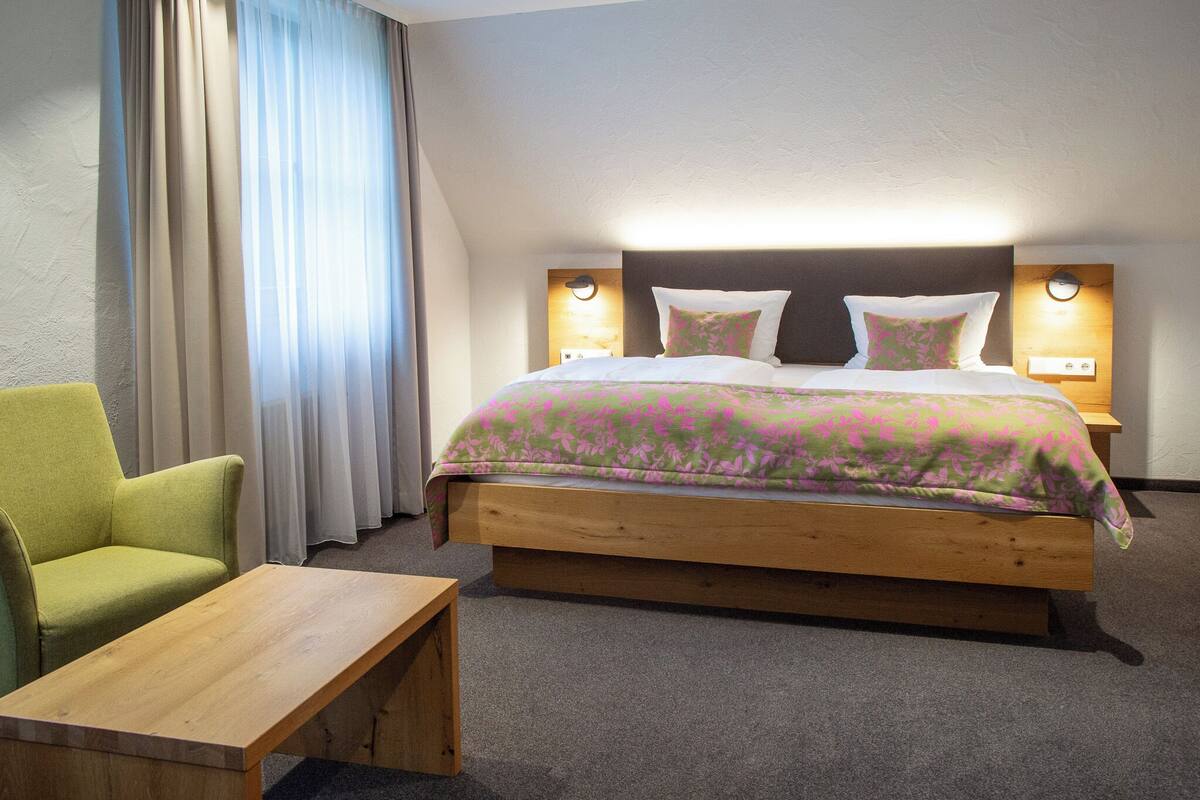 Ein Hotelzimmer mit einem hölzernen Bett, einem grünen Sessel, einem Nachttisch aus Holz und einer Wandleuchte.