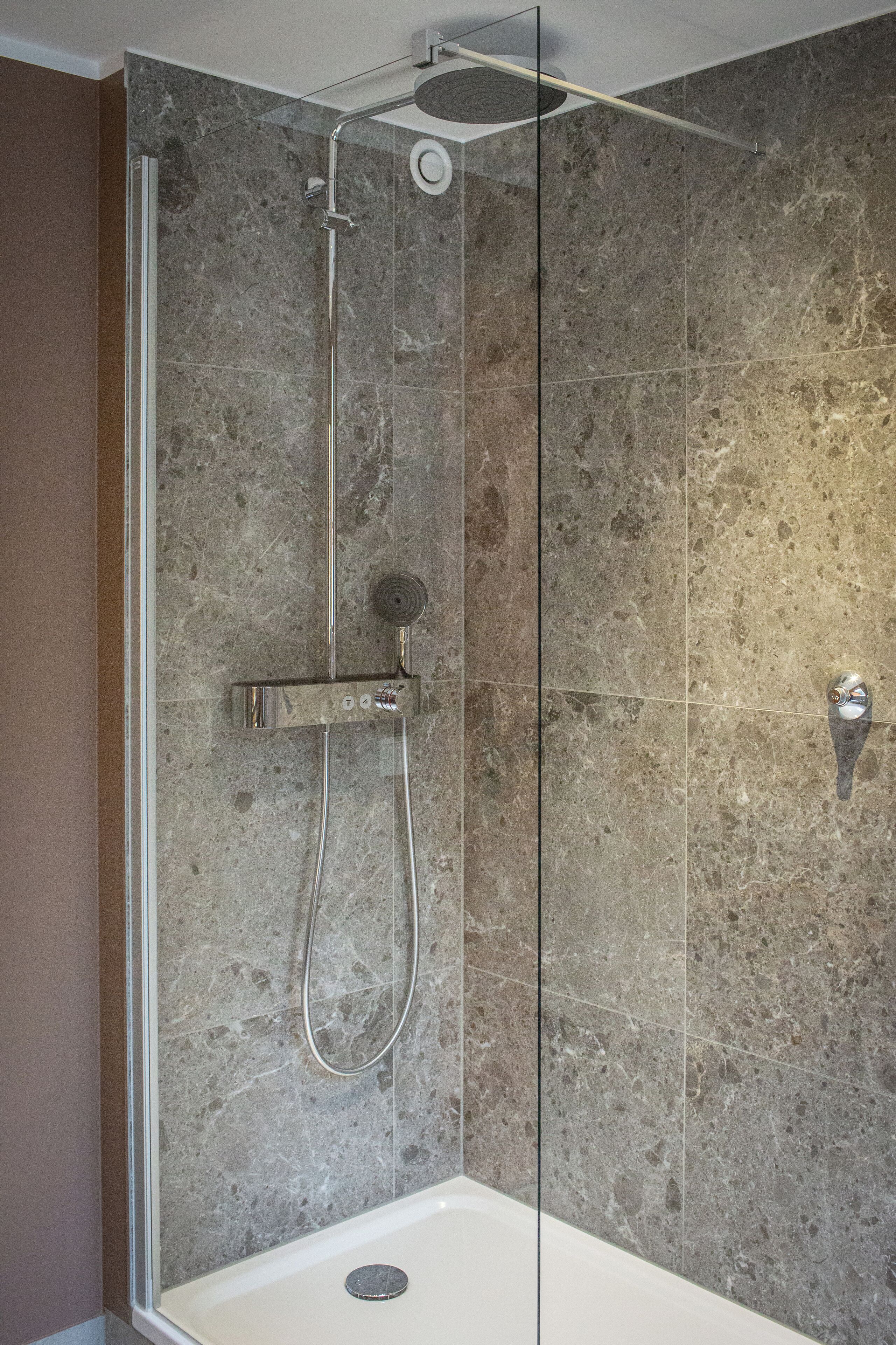 Une douche à l’italienne avec une porte en verre, une douchette à main et un panneau de commande fixé au mur.