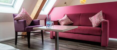 Una sala de estar moderna con un sofá rojo, un sillón púrpura y una mesa blanca. Hay dos botellas sobre la mesa, y una lámpara colgante la ilumina.