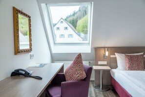 A hotel room with a desk, chair, bed, and a window with a view of a house and trees. - Hotel Hofgut Sternen | Ravennaschlucht (Breitnau)