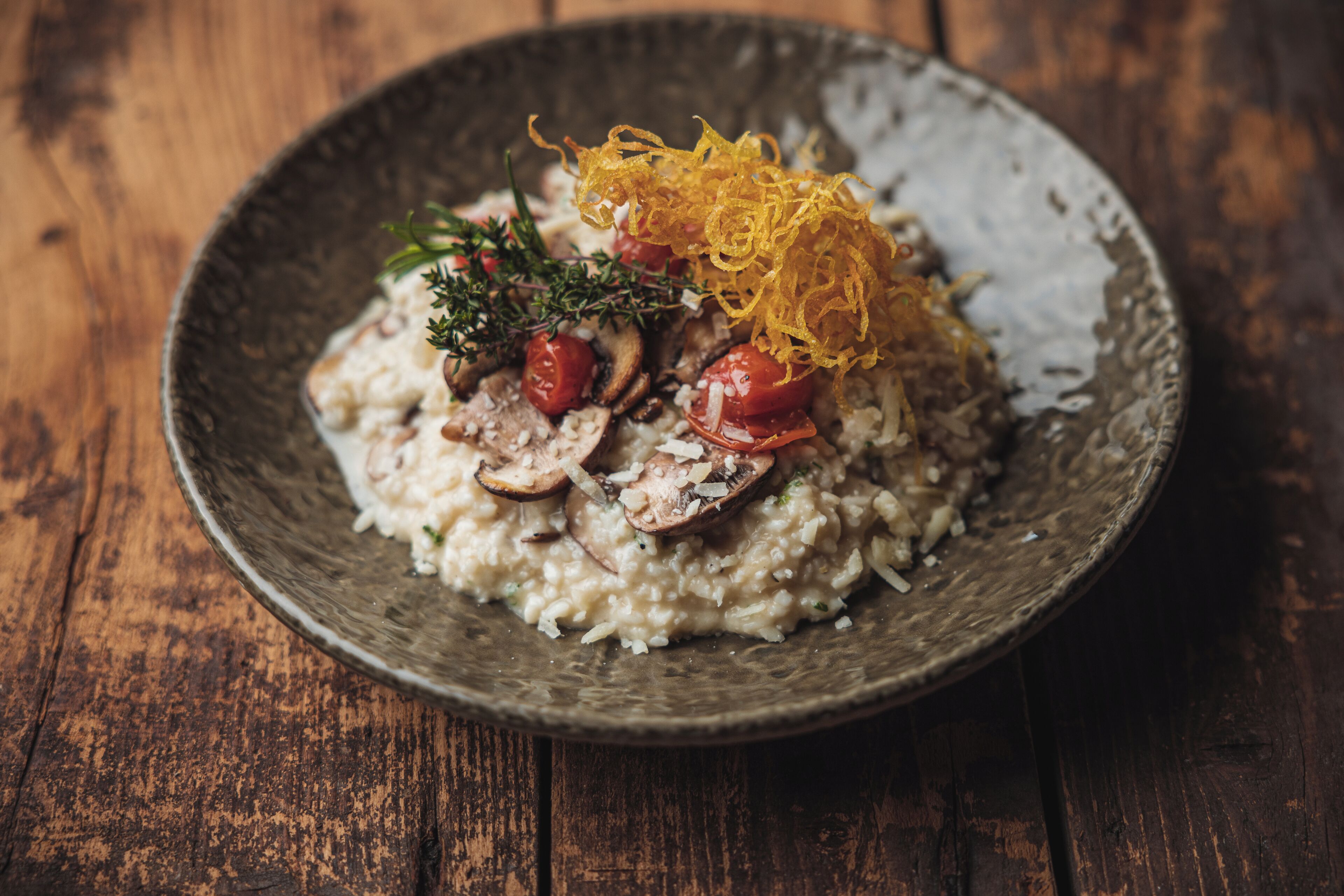 Una mesa de madera rústica con un bol texturizado que contiene un plato de risotto cubierto de champiñones, tomates cherry y hierbas, decorado con tiras crujientes.