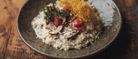 Una mesa de madera rústica con un bol texturizado que contiene un plato de risotto cubierto de champiñones, tomates cherry y hierbas, decorado con tiras crujientes.