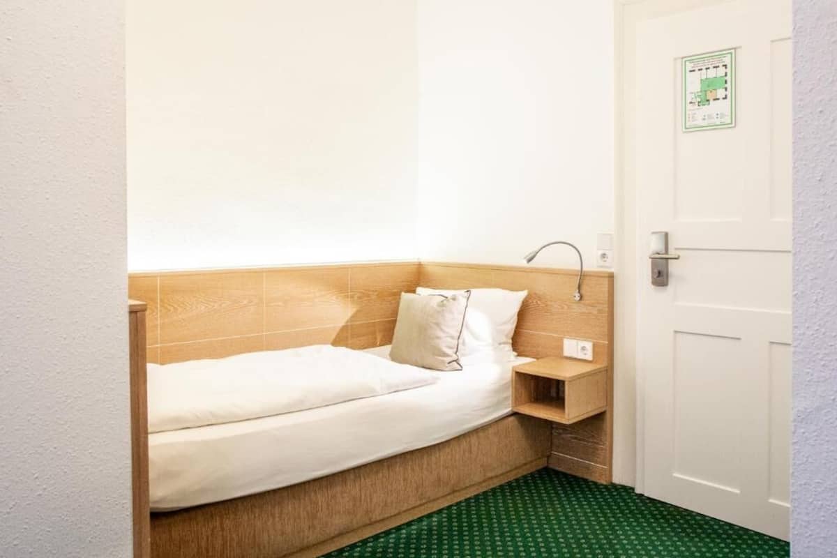Ein kleines, ordentlich eingerichtetes Schlafzimmer mit einem Einzelbett, einem hölzernen Kopfteil, einem Nachttisch und einem Teppich mit grünem Muster.