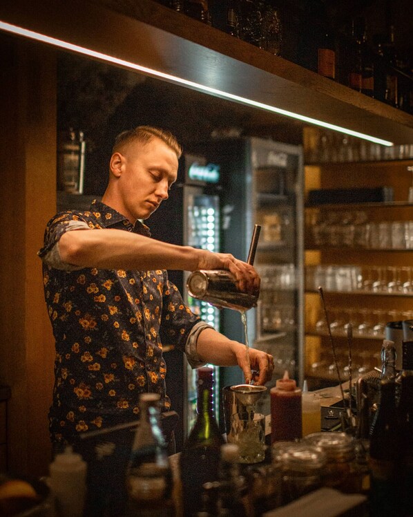 Um bartender preparando uma bebida num bar, com estantes cheias de garrafas e copos ao fundo.
