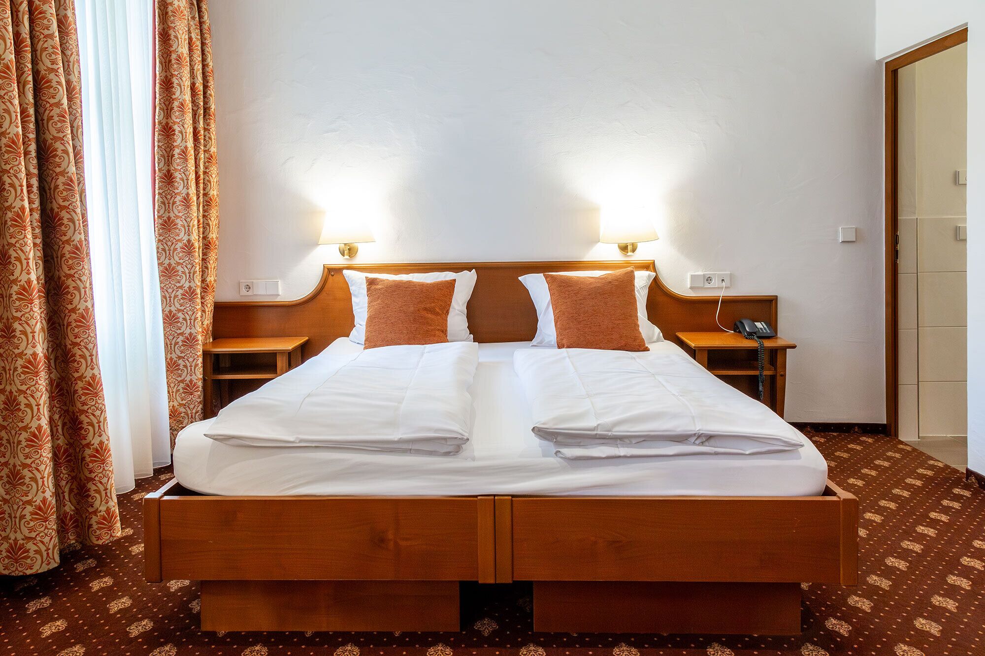 Cama doble con dos almohadas, cabecero de madera, mesitas de noche con lámparas y una alfombra estampada.