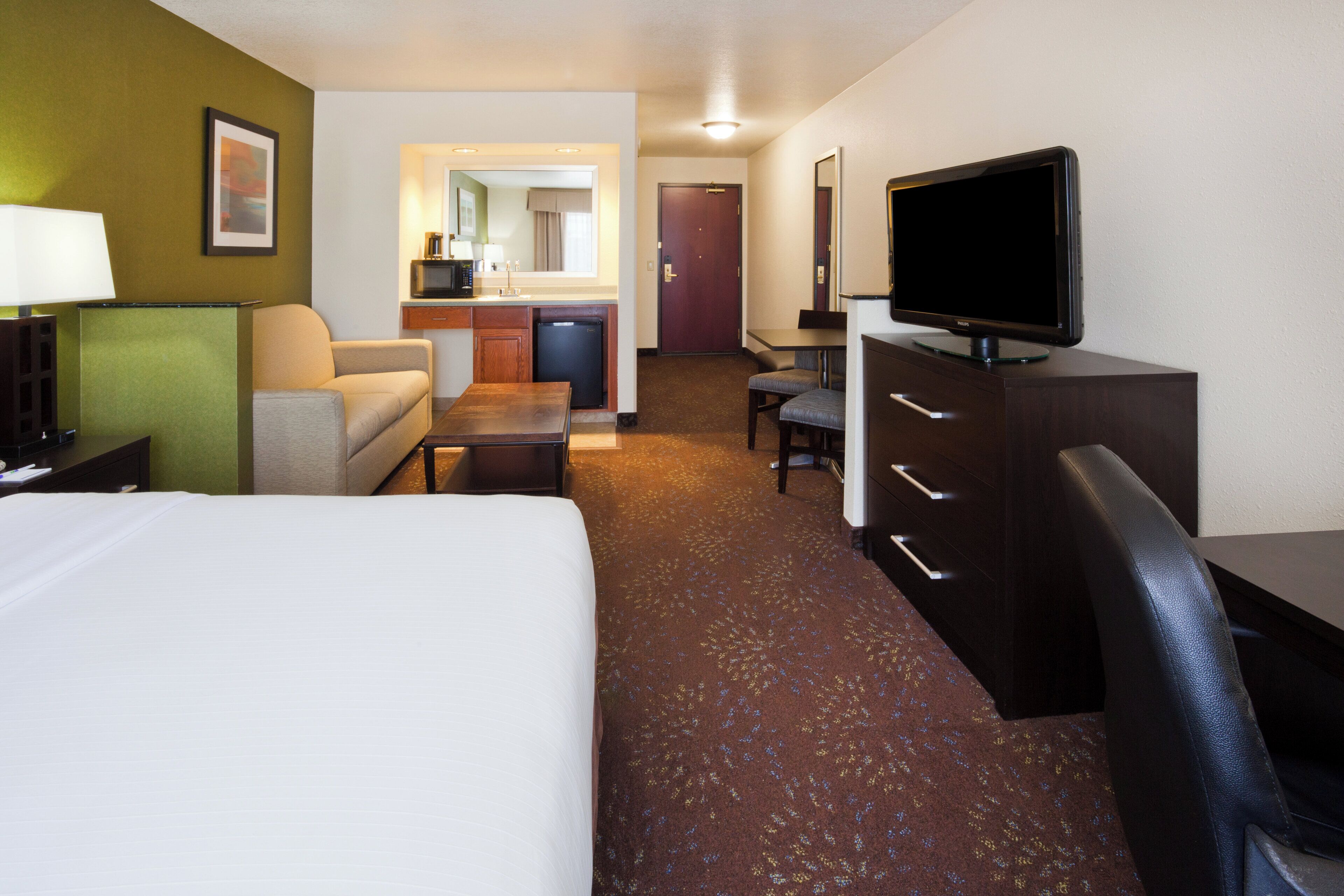 A hotel room with a bed, a desk, a television, a chair, a sofa, a small table, and a kitchenette area.
