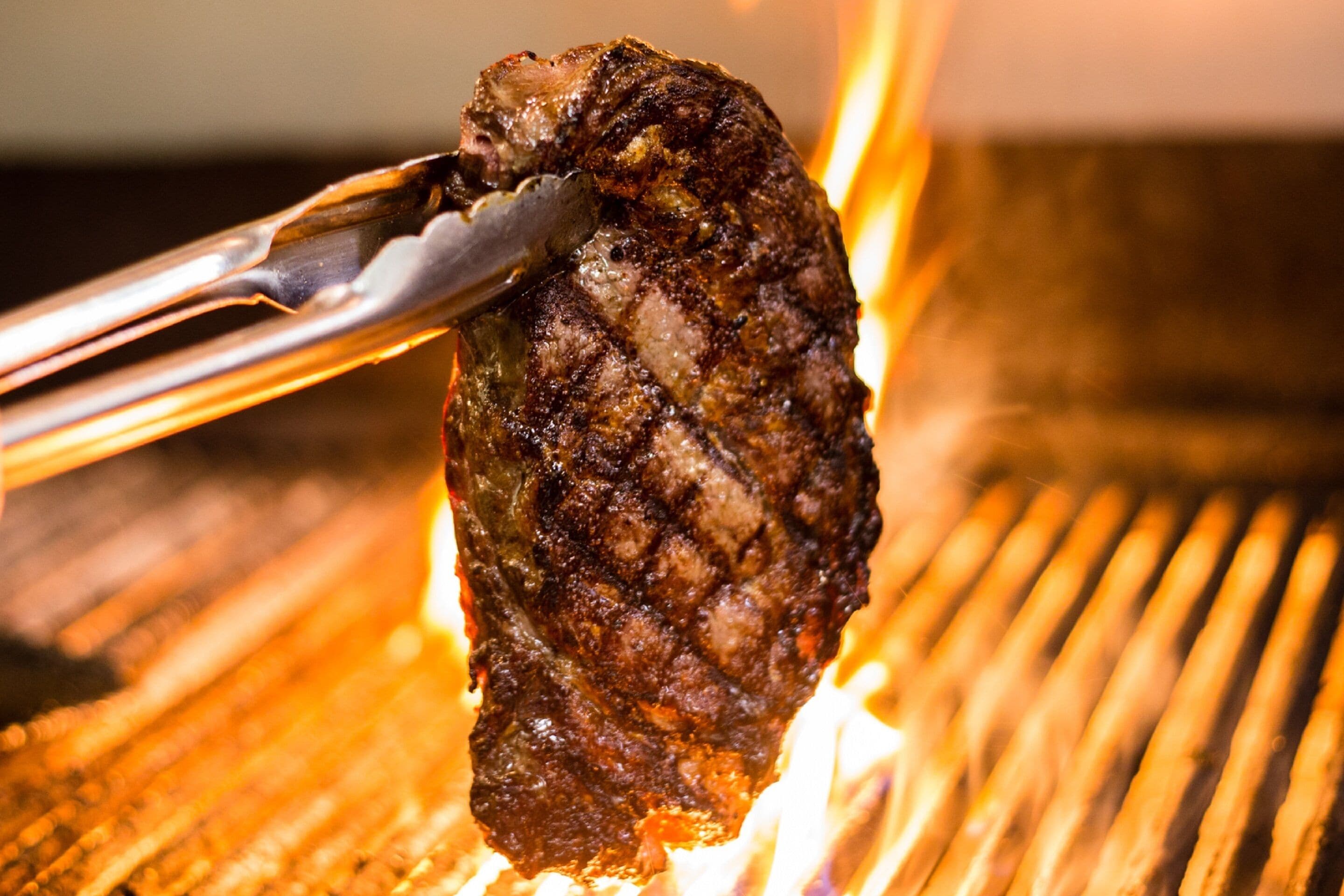 A close-up of a grilled steak being held by tongs over a grill.