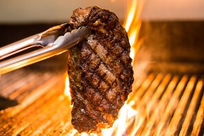 Gros plan d’un steak grillé, maintenu à l’aide de pinces au-dessus d’un gril.