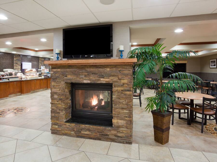 Eine Hotelhalle mit Kamin aus Stein, einem darüber montierten Fernseher, einer Frühstücksecke sowie mehreren Esstischen und Stühlen.