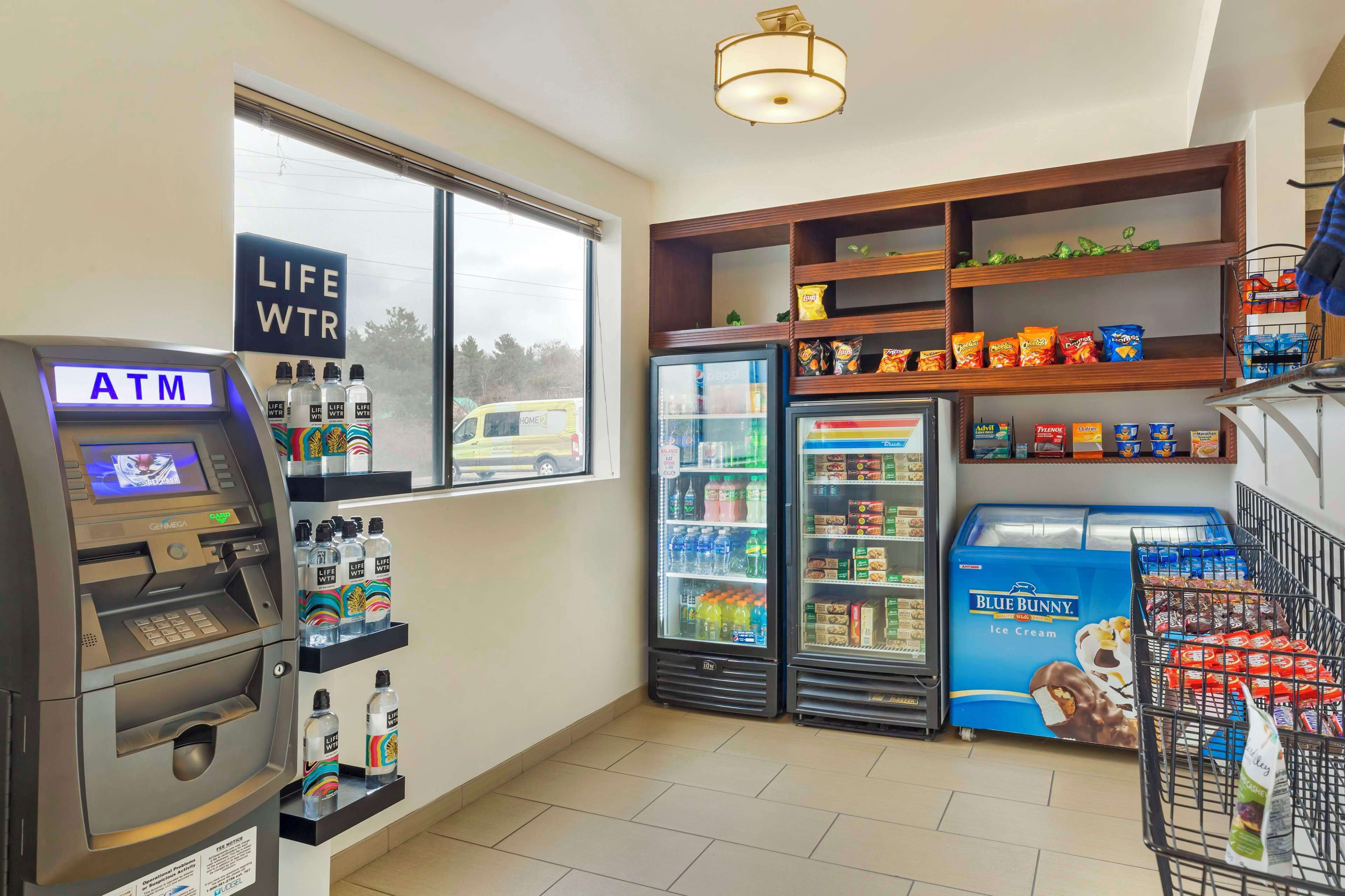 Un cajero automático, estantes refrigerados con bebidas y aperitivos, y un carrito de compras lleno de productos.