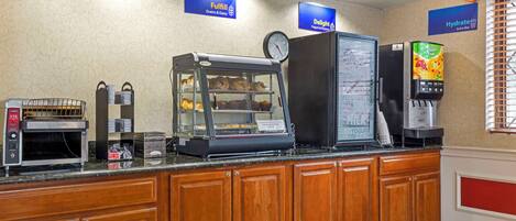 A breakfast bar with a coffee machine, a display case with pastries, a refrigerator, and a juice dispenser.
