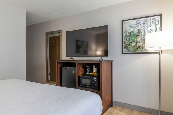 A hotel room with a bed, a television on a wooden stand, a microwave, a coffee maker, and a painting on the wall.