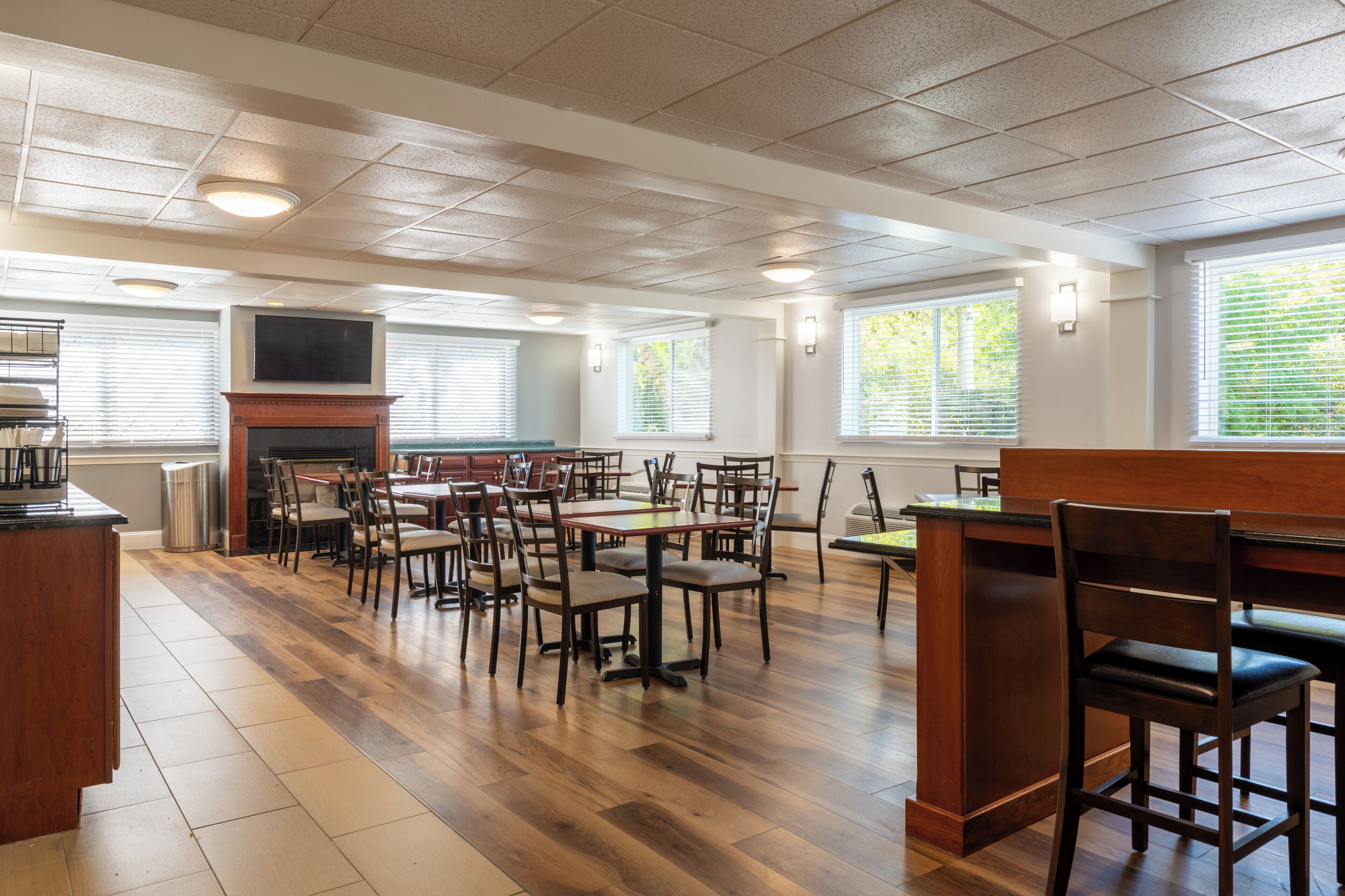 A spacious dining area with wooden flooring, multiple tables and chairs, a fireplace, and large windows with blinds.