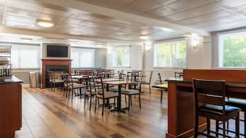 A spacious dining area with wooden flooring, multiple tables and chairs, a fireplace, and large windows with blinds.