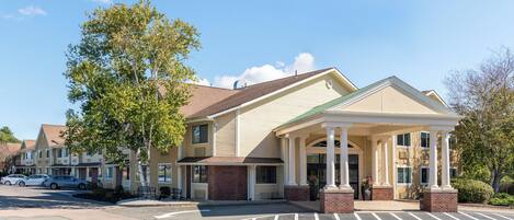 A multi-story hotel with a covered entrance, parking lot, and adjacent buildings.