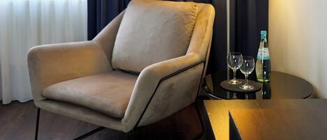 A modern hotel room with a beige armchair, a standing floor lamp, and a small table with two glasses and a bottle.