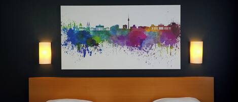 A hotel room with two beds, a city skyline artwork on the wall, and two wall-mounted lamps.