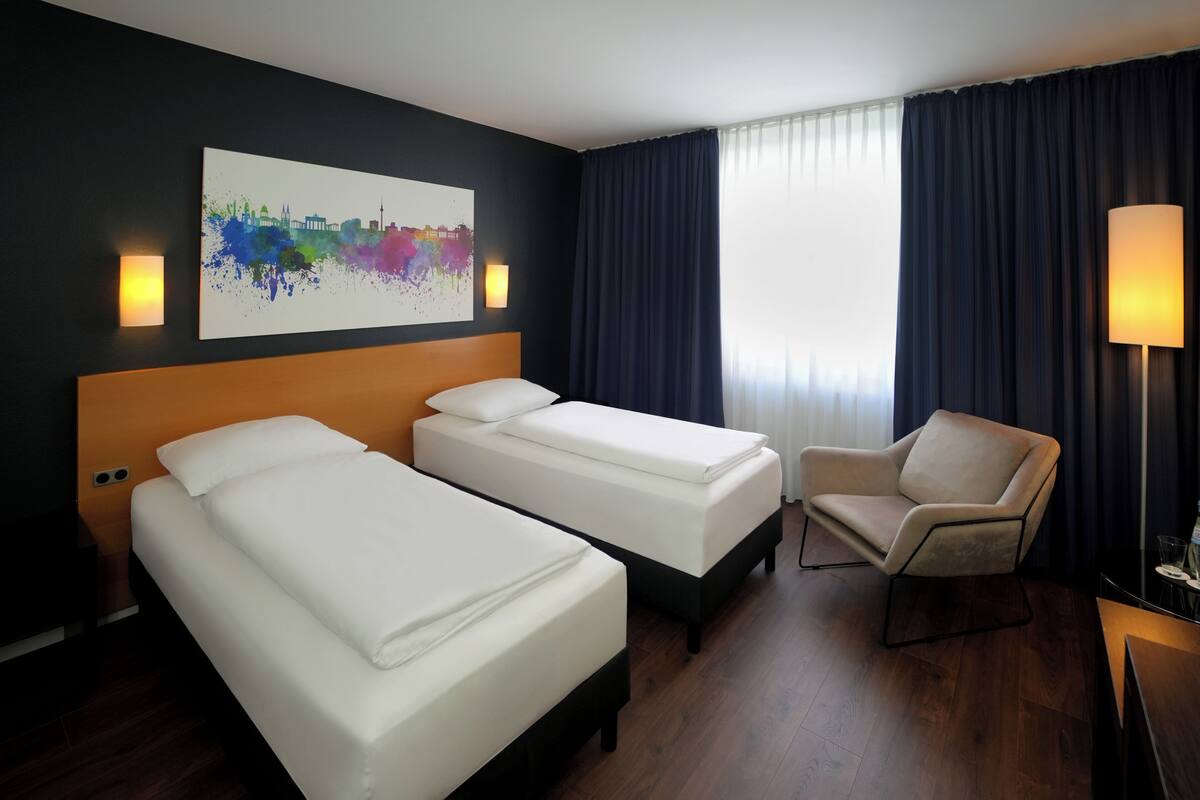 Quarto de hotel com duas camas, uma cadeira, um abajur e uma pintura da paisagem urbana na parede.
