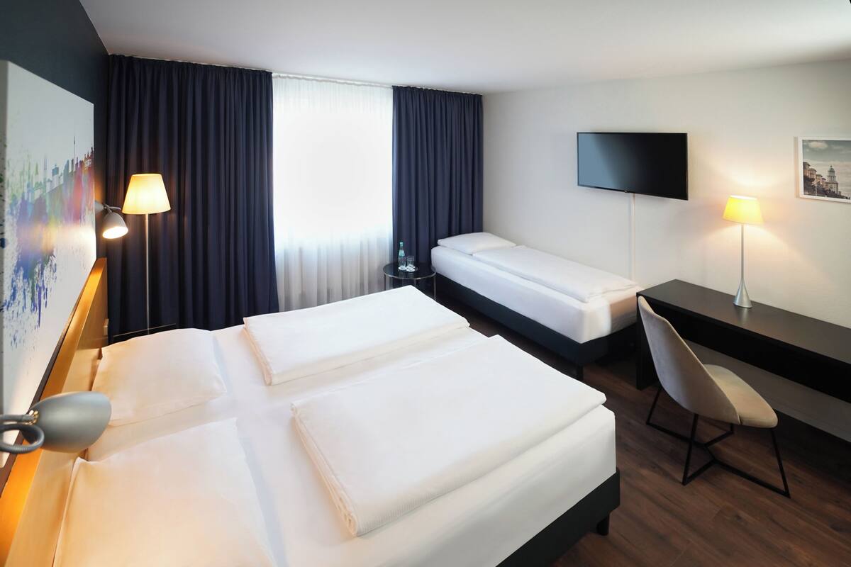 Quarto de hotel com duas camas, uma escrivaninha, uma cadeira, uma televisão e um quadro na parede.