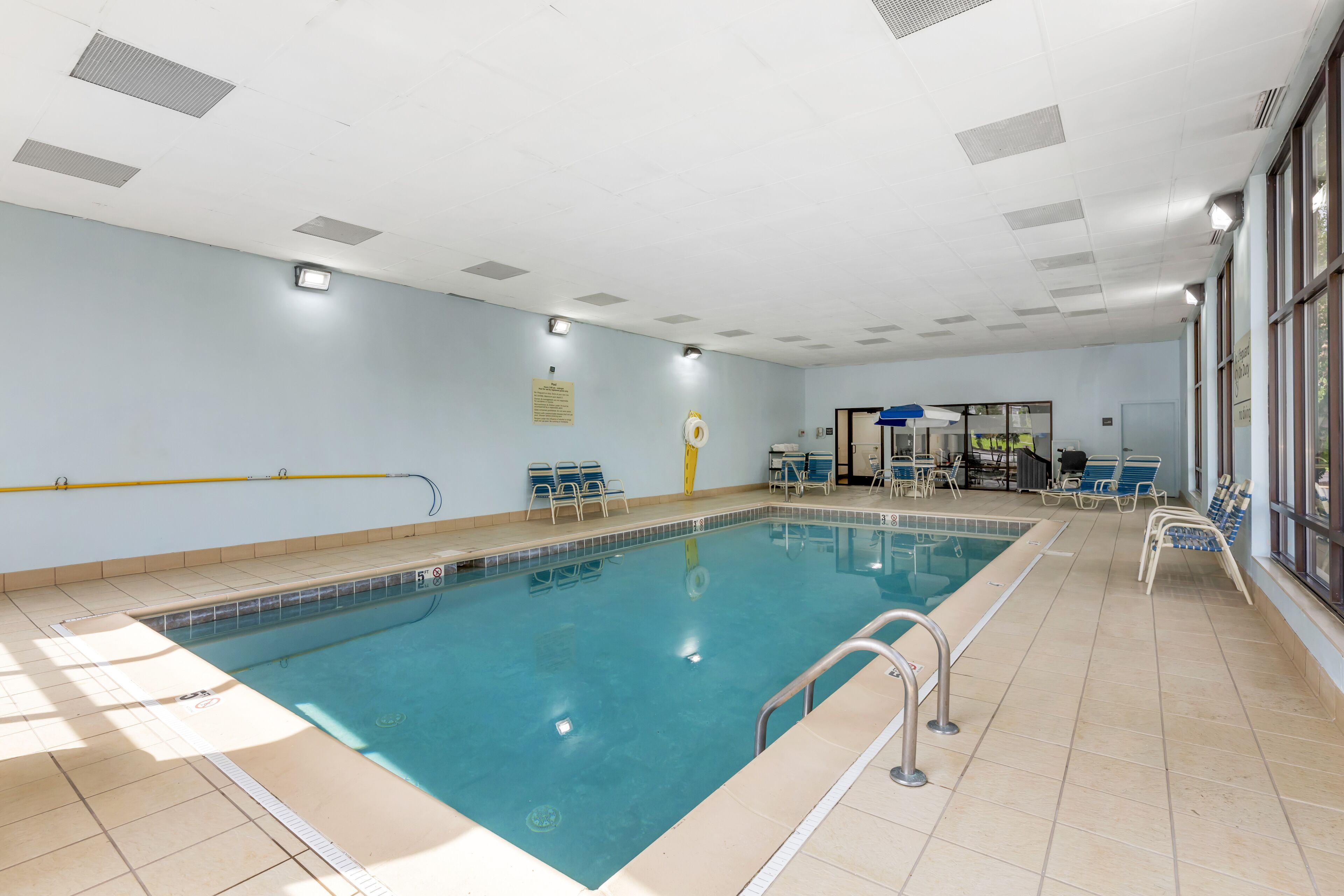 A swimming pool with a tiled deck, sunlit by natural light, and a seating area with chairs and a table.