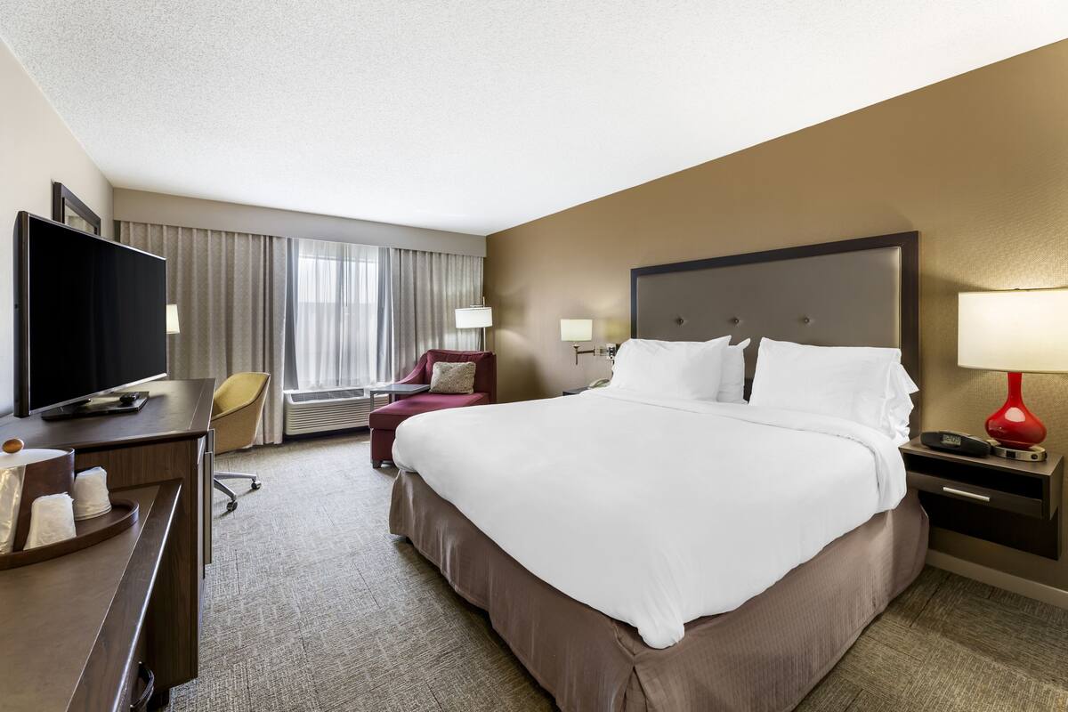 A hotel room with a large bed, a desk with a flat-screen TV, a chair, and a window with curtains.