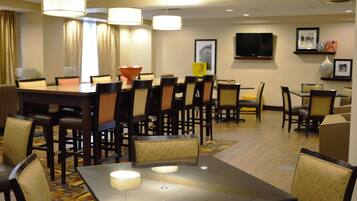A hotel dining area with multiple tables and chairs, a television on the wall, and a patterned carpet.