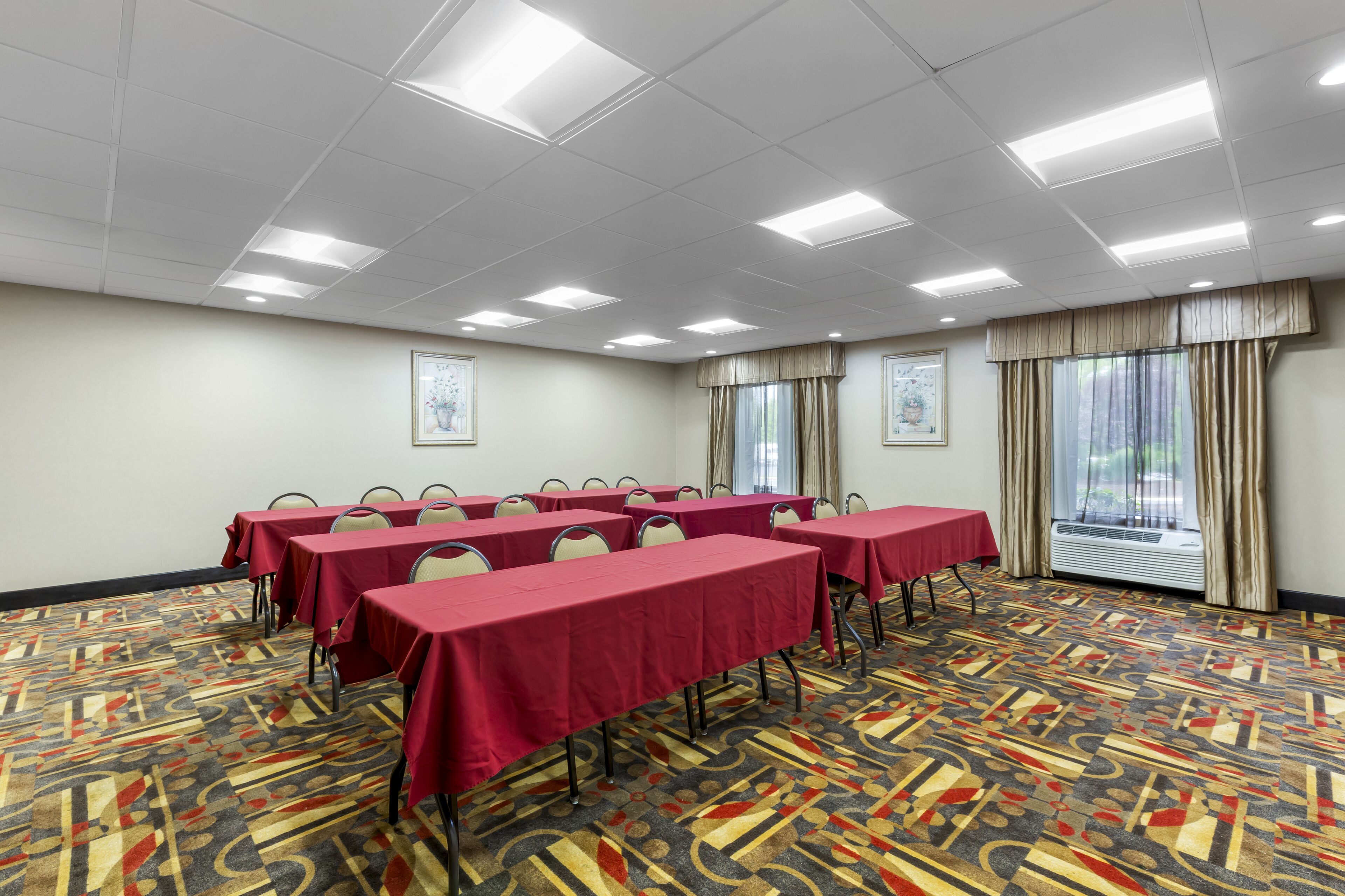 A conference room with tables covered in red tablecloths, arranged in rows, with chairs at each table. The room has a patterned carpet, white walls, and a large window with curtains.