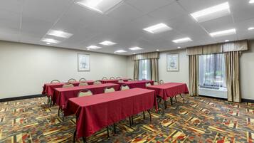 A conference room with tables covered in red tablecloths, arranged in rows, with chairs at each table. The room has a patterned carpet, white walls, and a large window with curtains.
