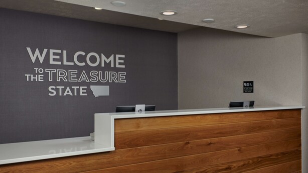 A reception desk with a grey wall behind it displaying "WELCOME TO THE TREASURE STATE" and a wooden panel design.
