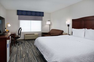 Una habitación de hotel con una cama grande, un escritorio con computadora, una silla, una ventana con persianas y una lámpara fijada a la pared.