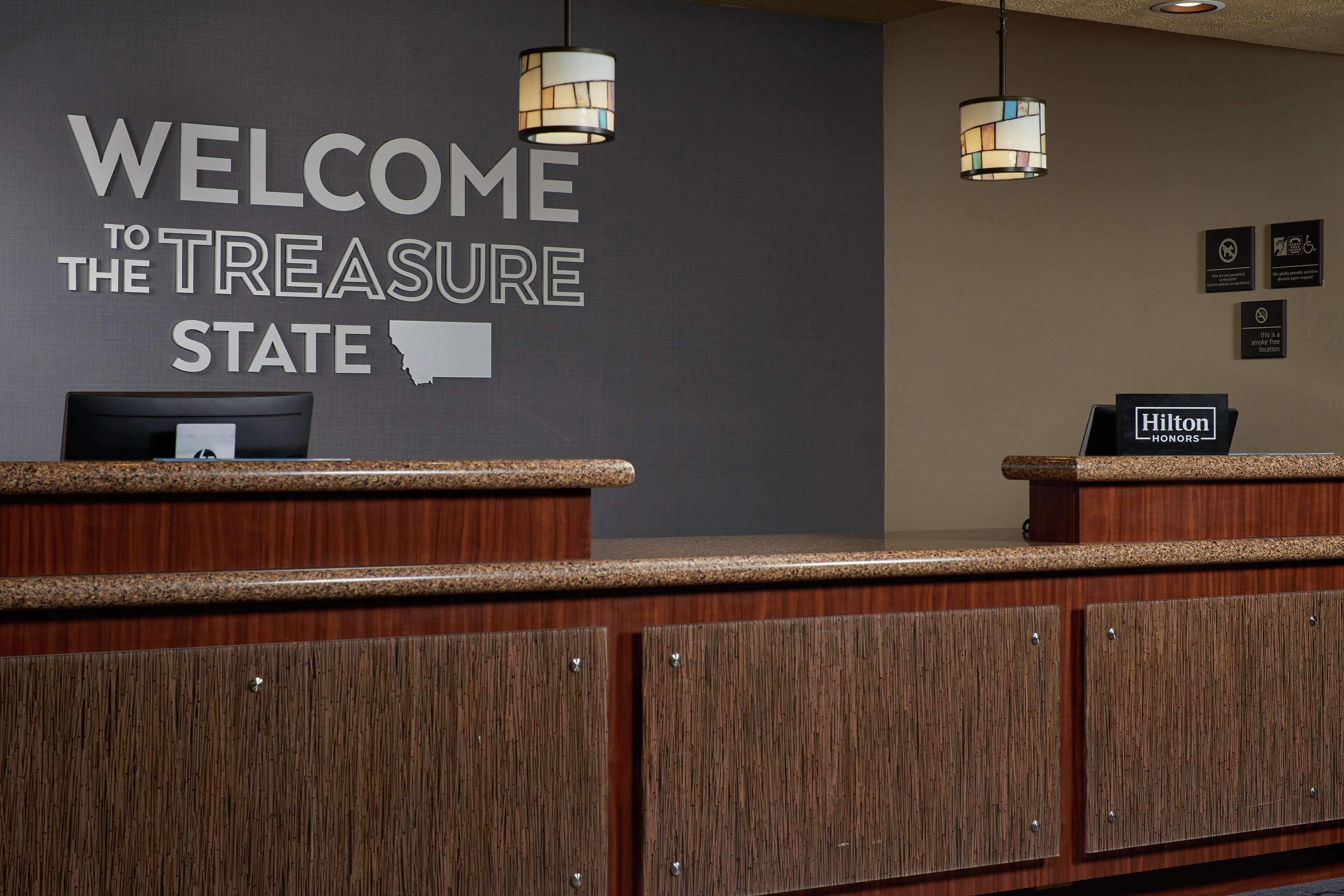 Un mostrador de recepción con un cartel que dice "Welcome to the Treasure State", un cartel de Hilton Honors y una foto enmarcada sobre el mostrador.
