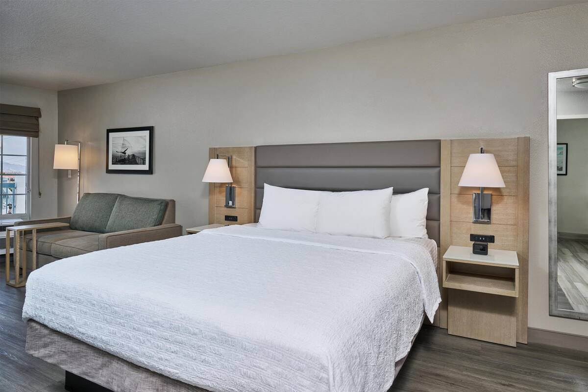 A hotel room with a large bed, two bedside tables with lamps, a chair, a small table, and a framed picture on the wall.