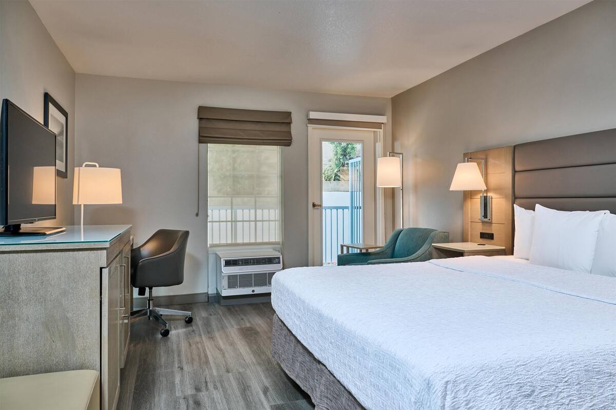 A hotel room with a bed, desk, chair, and a window with blinds.