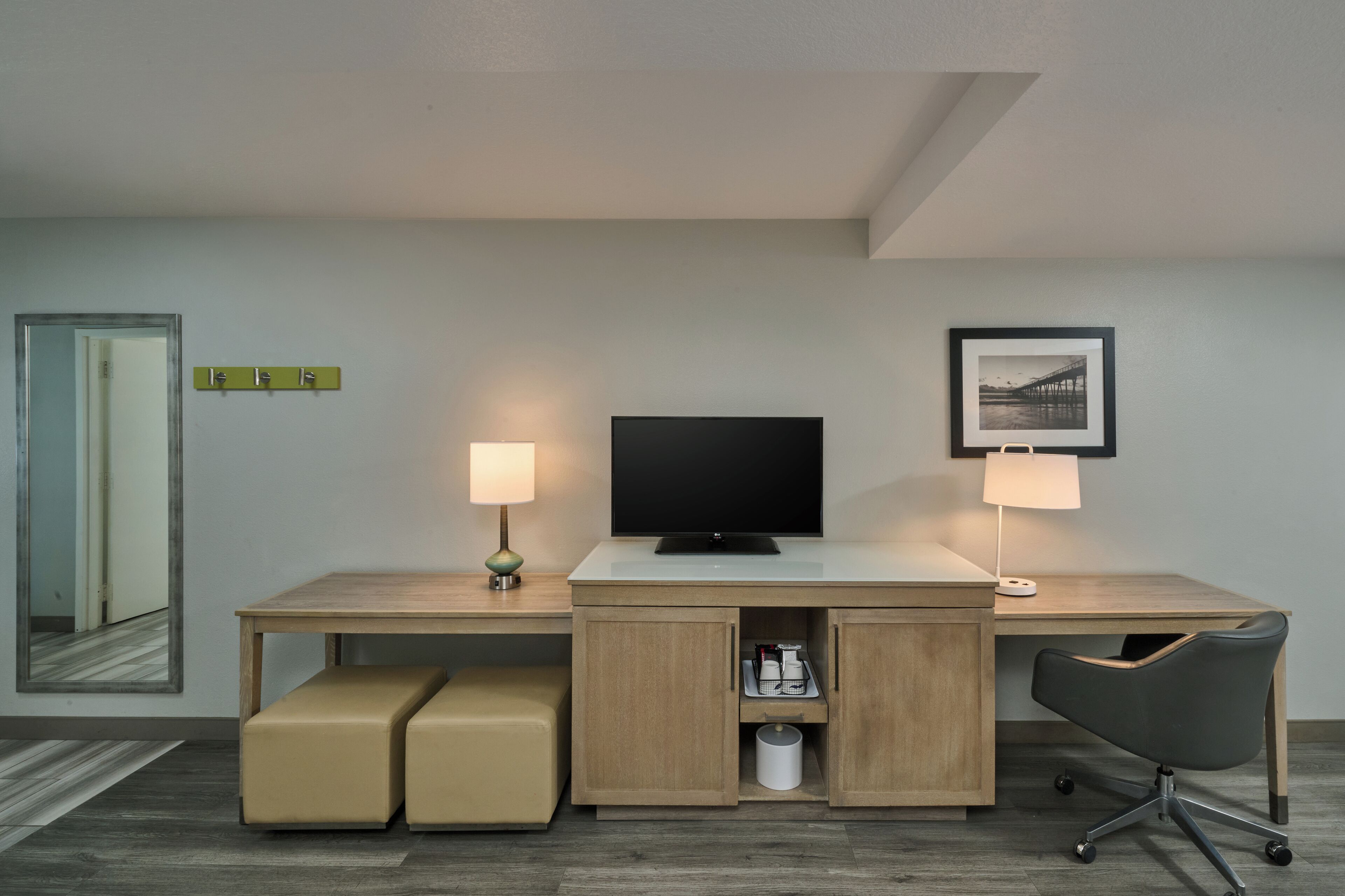 A hotel room with a wooden desk, a television, two bedside lamps, a framed picture on the wall, and a mirror.