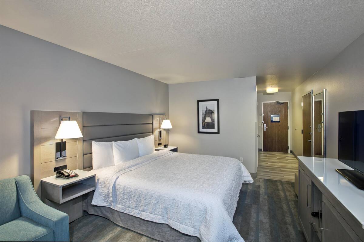 A hotel room with a bed, bedside lamps, a chair, a desk with a telephone, a television, and a framed picture on the wall.