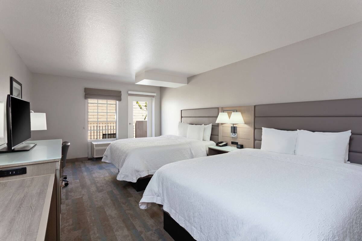 A hotel room with two beds, a TV, a desk, and a window with blinds.