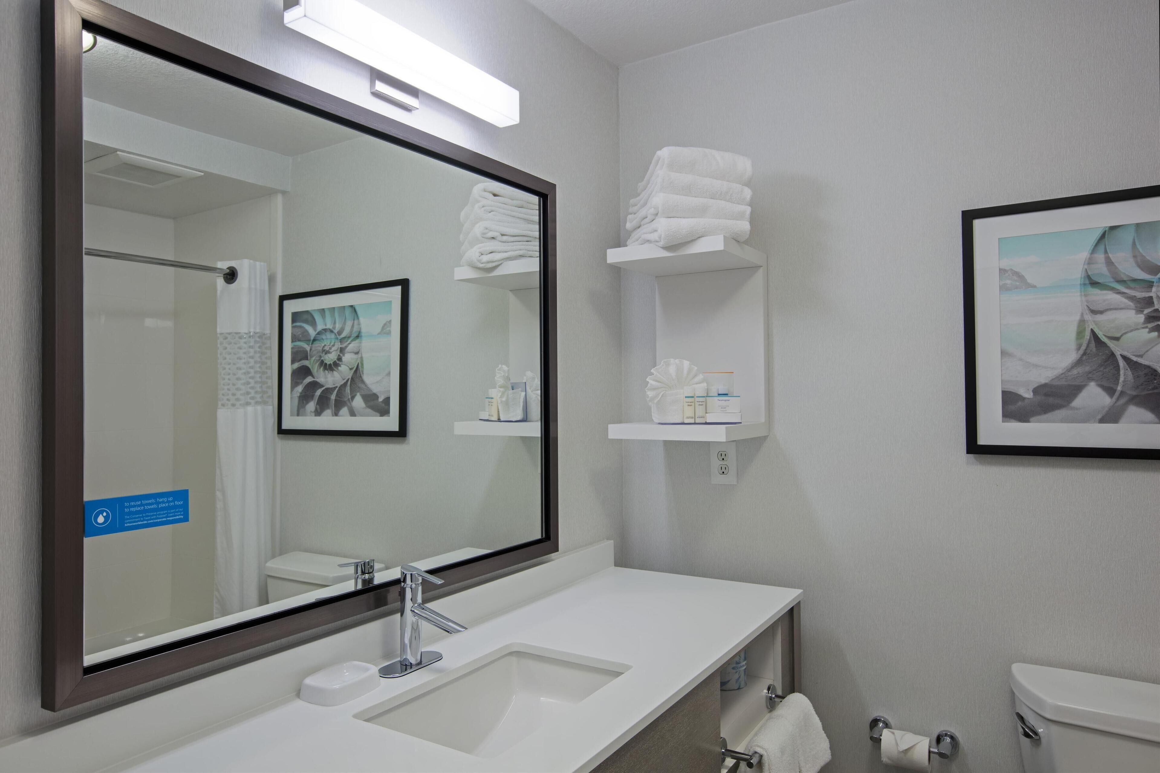 A modern bathroom with a large mirror, white sink, and toilet. There are shelves with towels and toiletries.