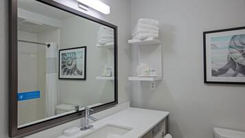A modern bathroom with a large mirror, white sink, and toilet. There are shelves with towels and toiletries.