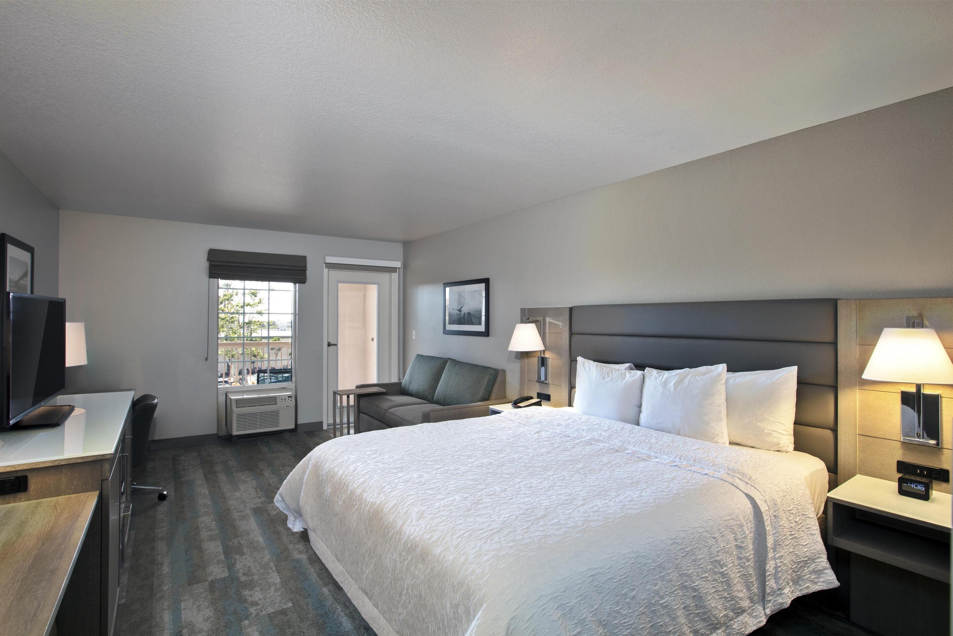 A hotel room with a large bed, a desk with a computer, a chair, a TV, and a window with a view of the outdoors.